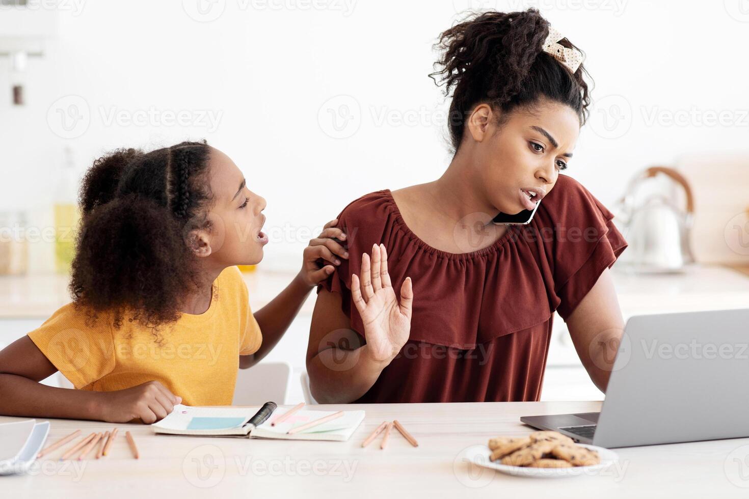 Angry african american teen girl sitting by working mom, asking for attention, kitchen interior. Busy black lady working from home, using laptop and having phone conversation, have no time for kid photo