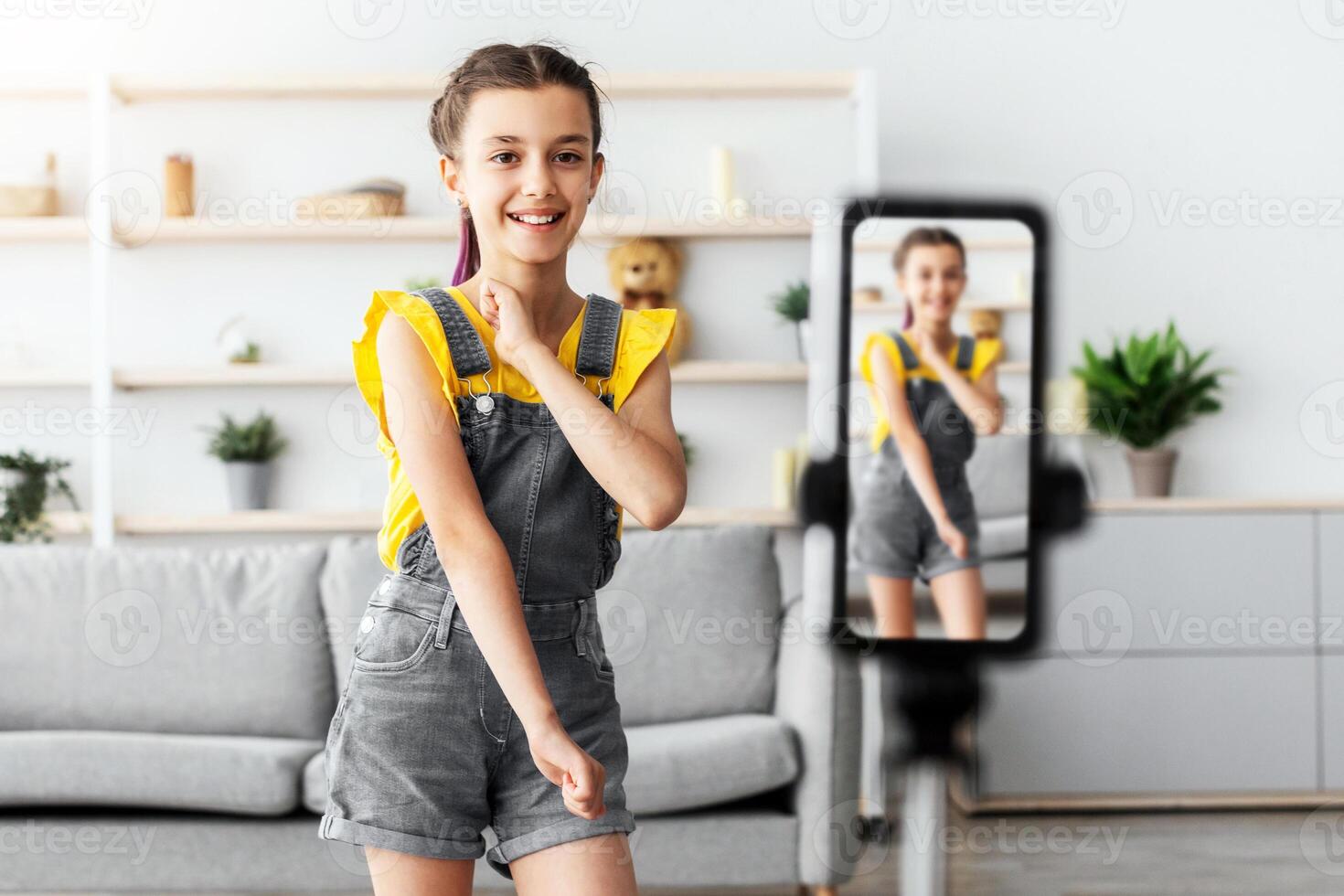 Modern Content Creator Concept. Portrait of positive teenage girl filming her dancing tutorial for social network, using cellphone on tripod in living room, selective focus photo