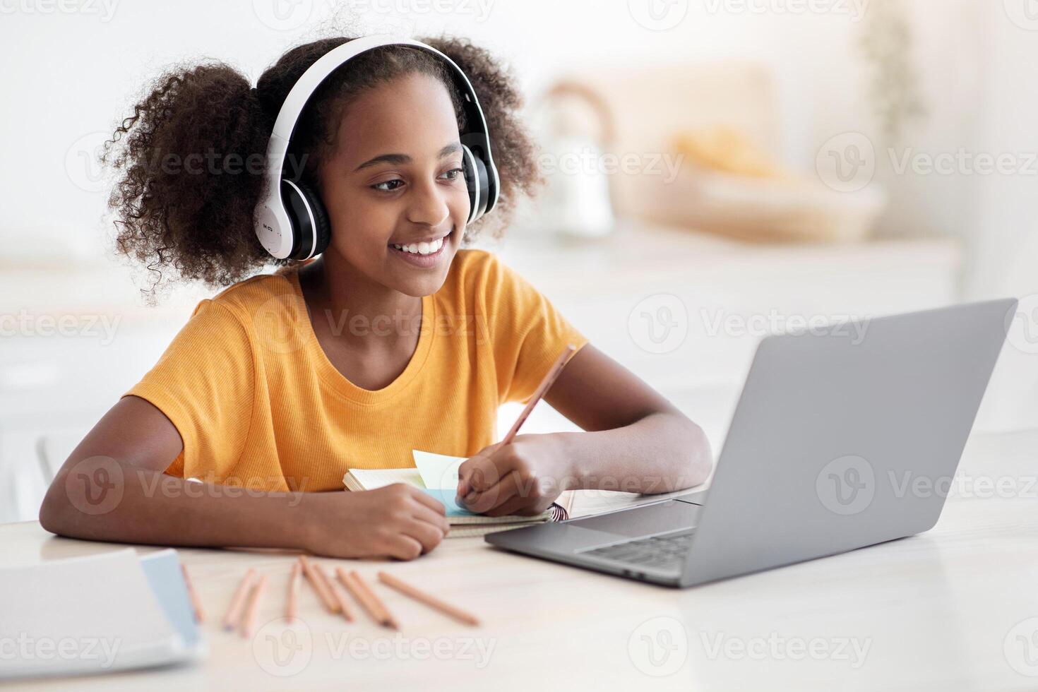 Cheerful black girl studying online, using laptop and headset at home, copy space. Cute african american teen kid having online lesson, drawing, kitchen interior. Home schooling concept photo