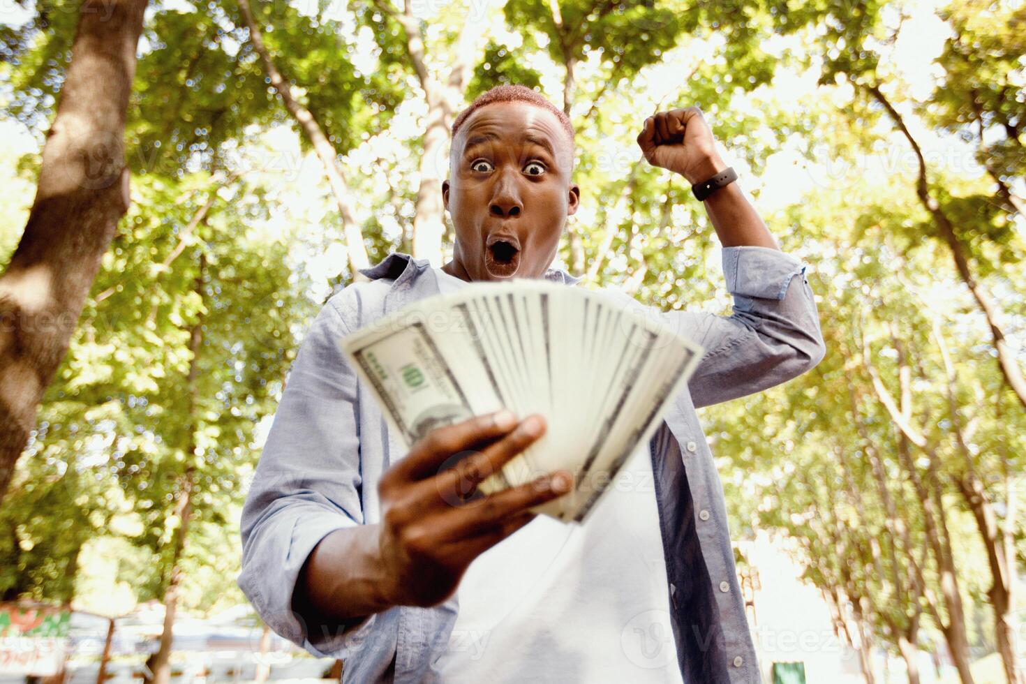 Big win concept. Excited African American guy holding fan of American money at park. Triumphant black man celebrating increase in profit, winning lottery, gaining unexpected wealth photo