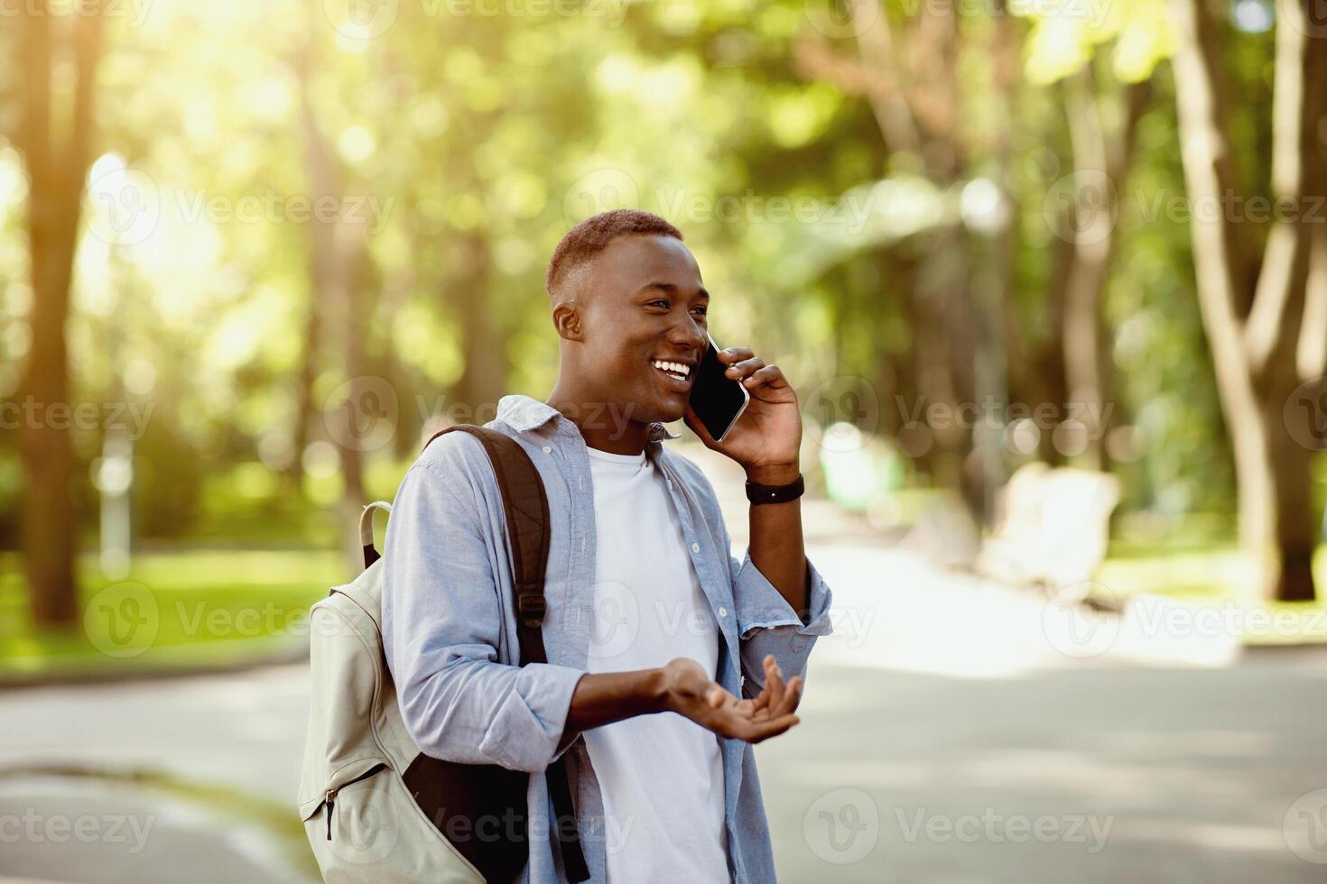 Happy black guy with backpack speaking on mobile phone at city park, copy space photo