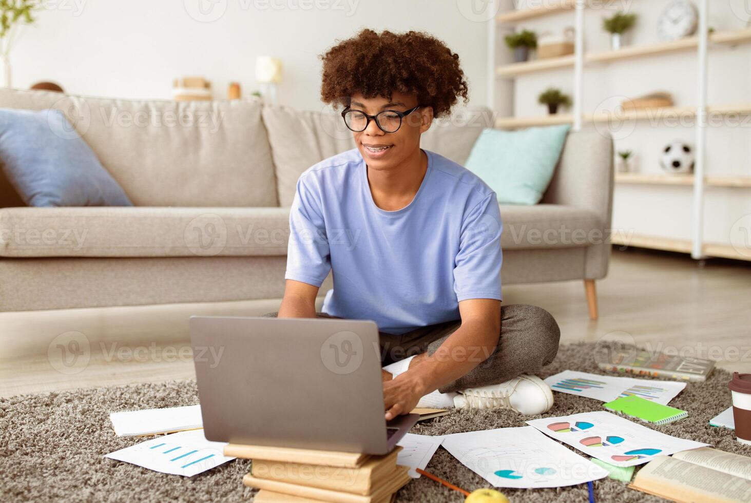 Home education concept. Black teen student working on study project, using laptop on floor indoors, full length. African American adolescent with scattered papers getting ready for online exam photo