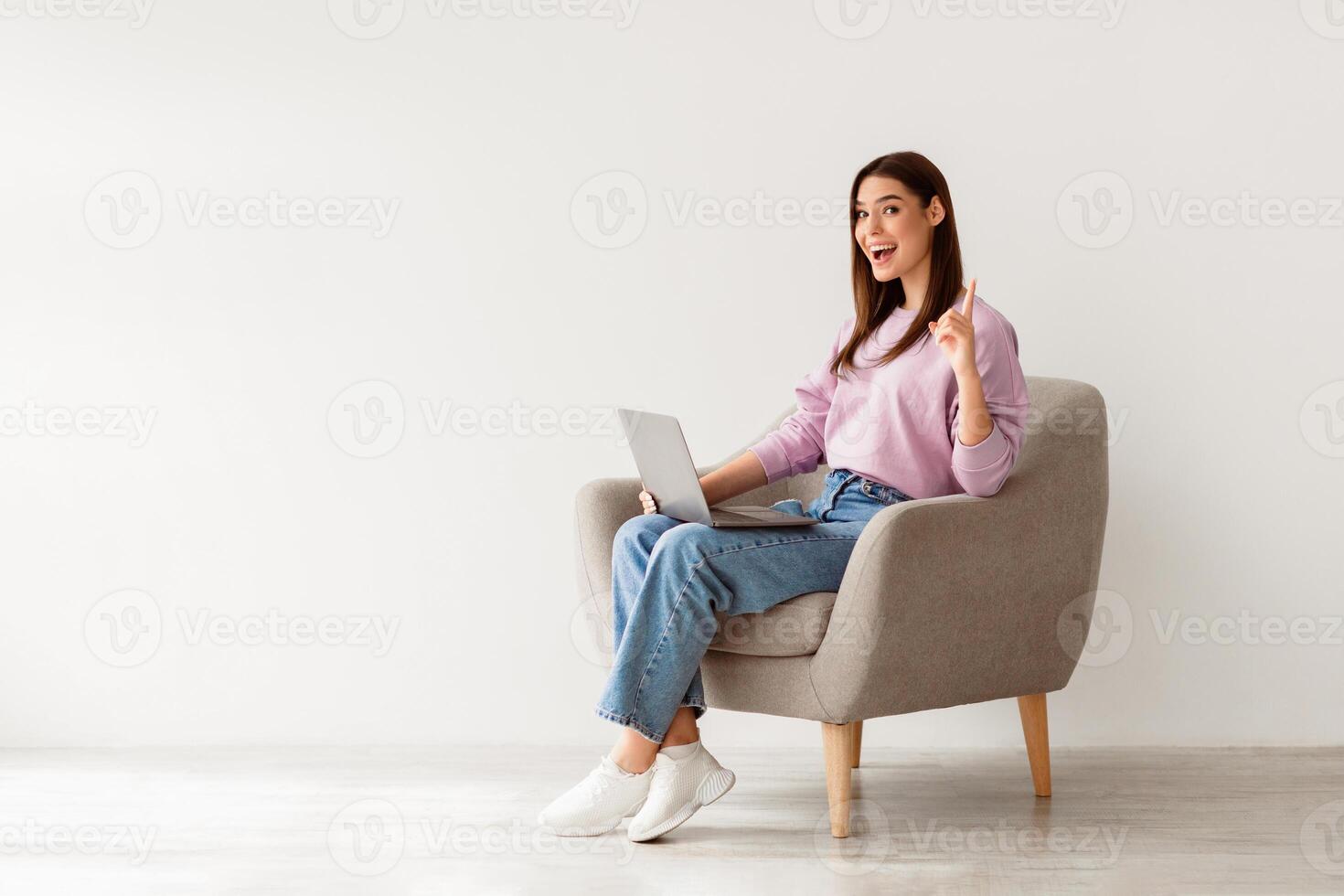 Millennial Caucasian woman having creative idea, gesturing eureka, sitting in armchair with laptop pc against white studio wall, copy space. Young lady finding solution to problem photo
