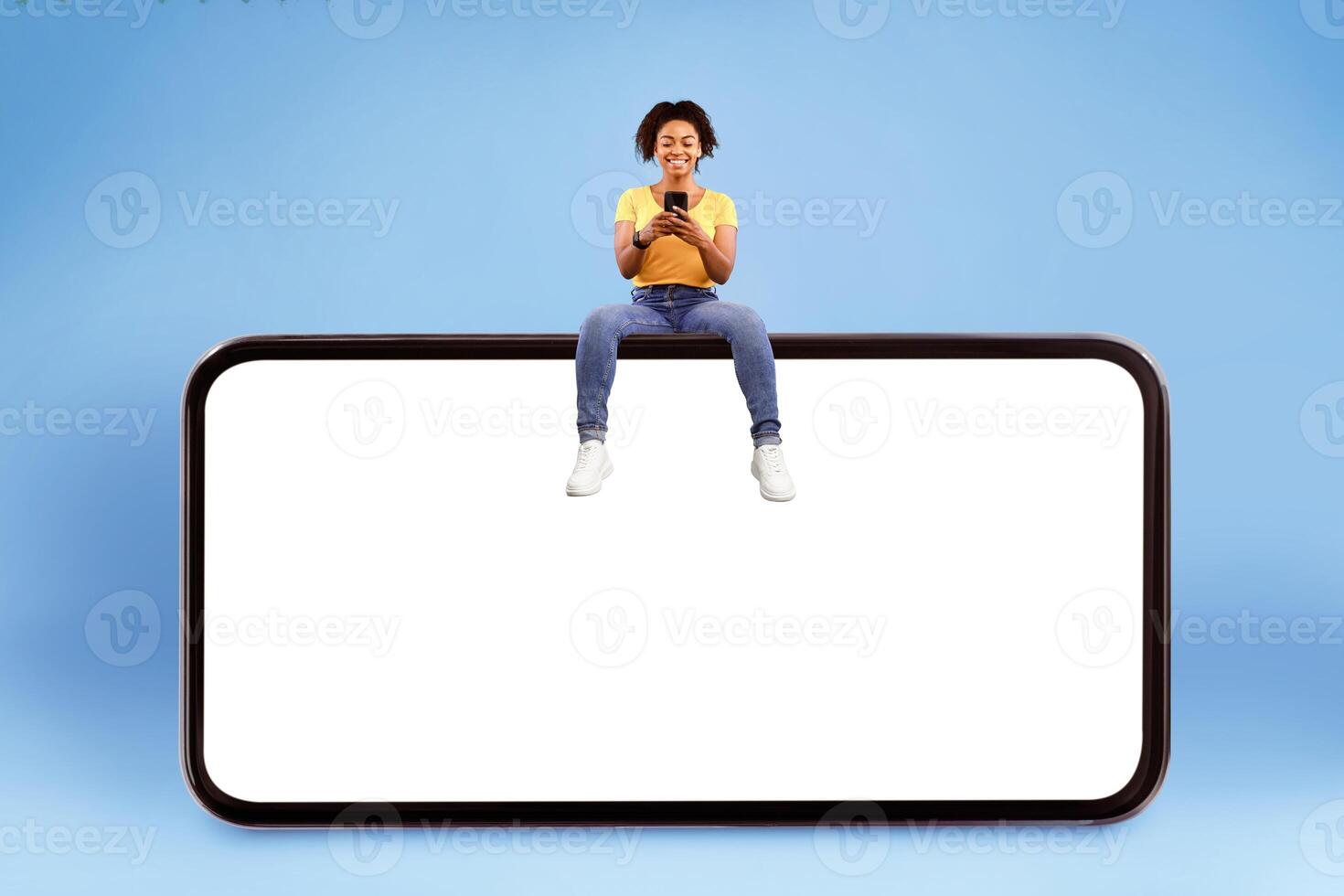 Cheerful young African American lady sitting on big smartphone with empty screen, using mobile device, demonstrating mockup for website, advertisement or app over blue studio background photo