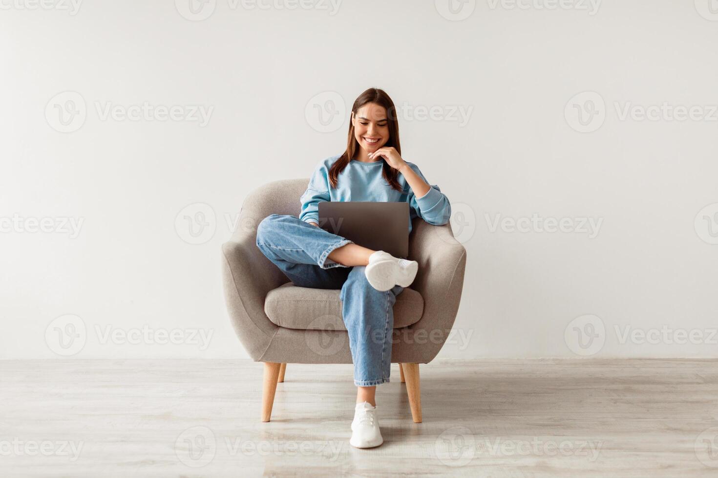 Millennial Caucasian lady in casual wear using laptop, sitting in armchair, having online conference against white studio wall. Young woman communicating online, taking part in webinar photo