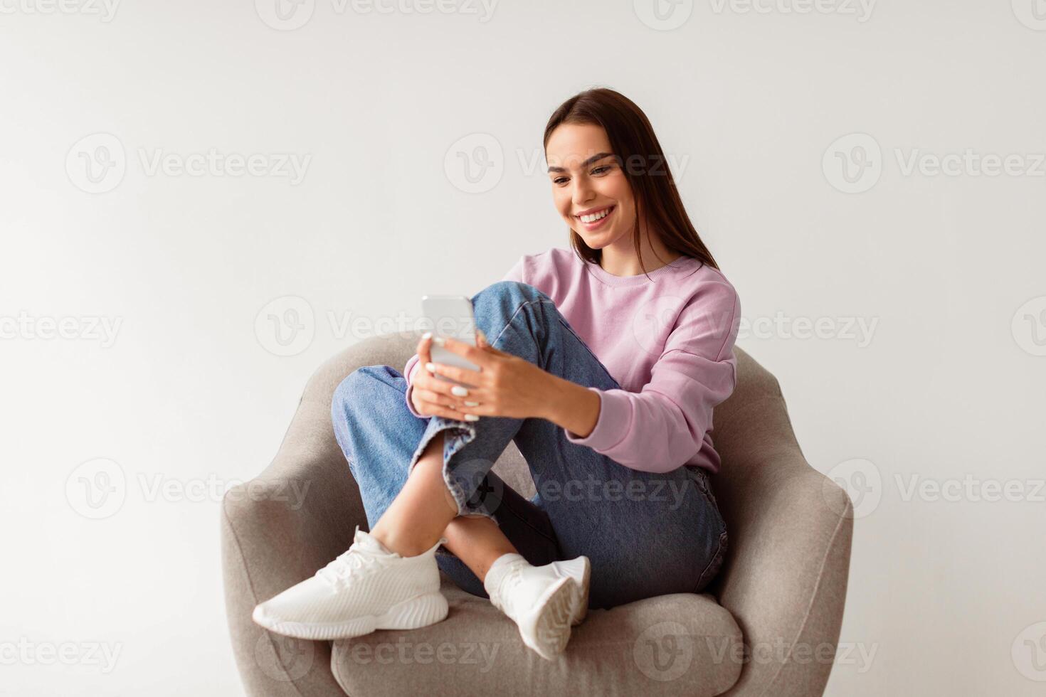 Full length of cheery millennial woman sitting in armchair with crossed legs, using smartphone, writing sms message, chatting with friend, browsing internet, watching online on white background photo