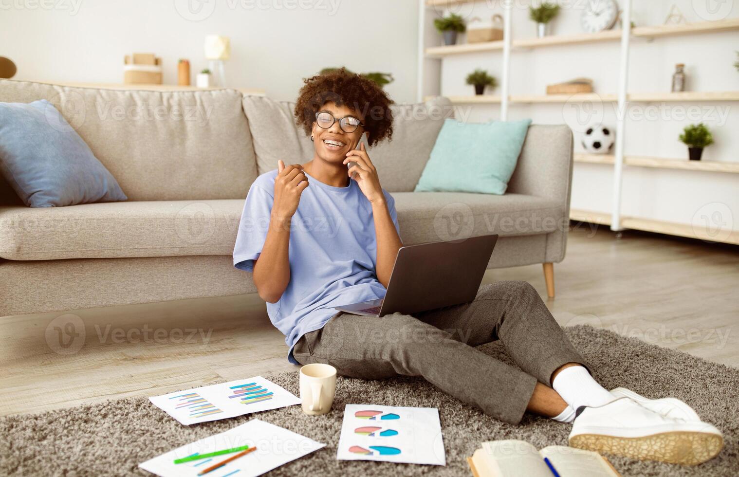 Happy black teenager sitting on floor with laptop and study materials, speaking on smartphone at home. Remote education, distance communication during covid lockdown concept photo