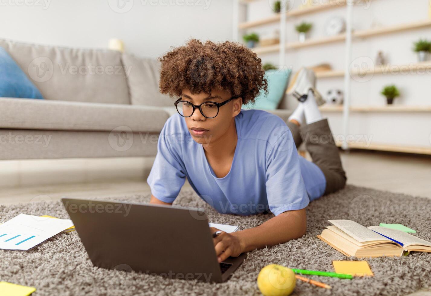 Focused black teenager using laptop pc for remote education, lying on floor and typing on keyboard at home. African American adolescent participating in webinar or online class indoors photo
