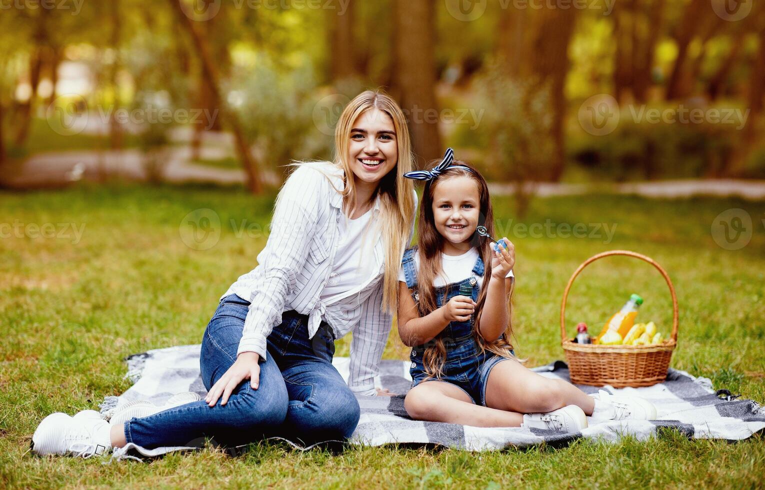 Family Picnic. Mother And Daughter Blowing Soap Bubbles Sitting On Plaid In Park Outdoors, Smiling To Camera photo