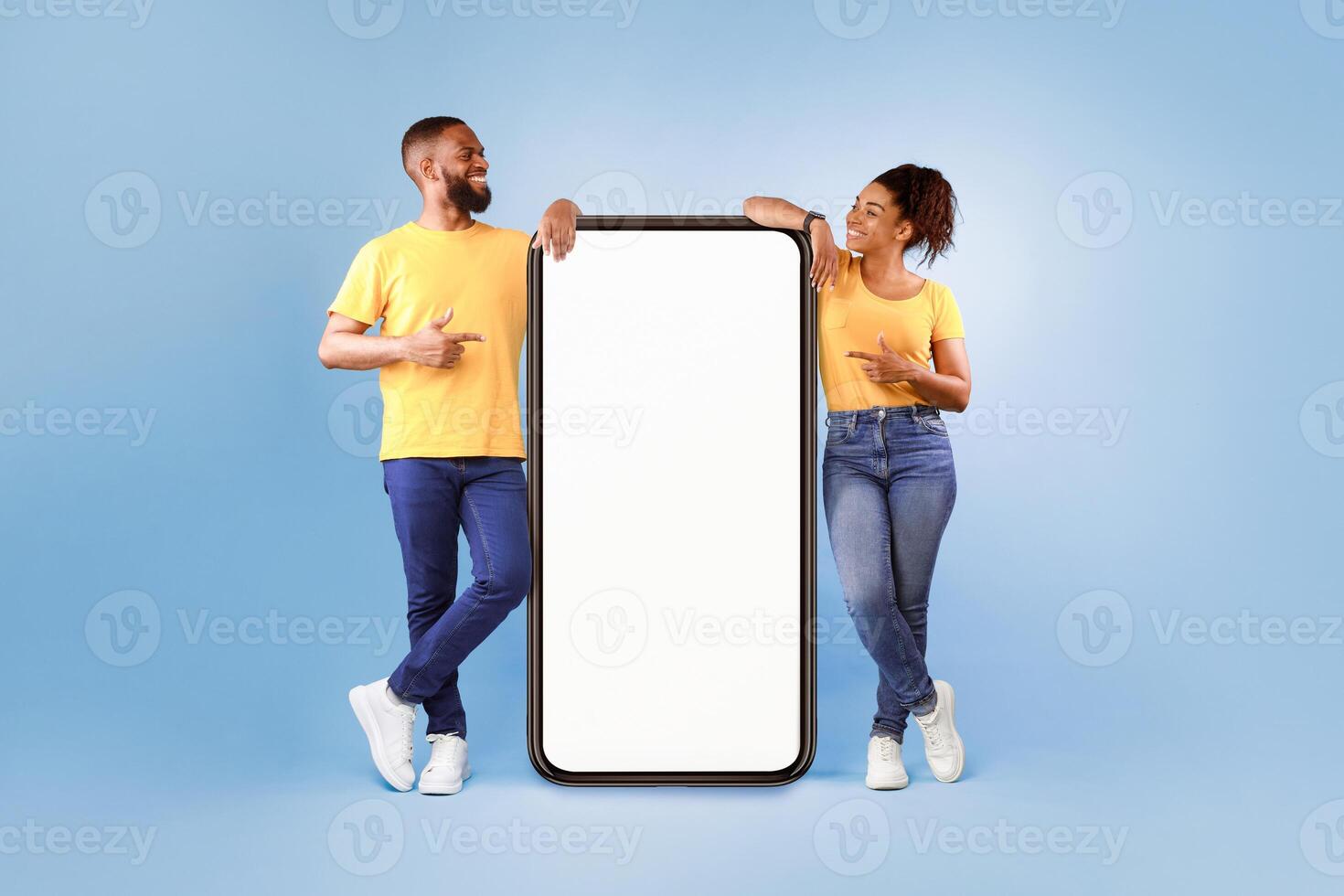 Hot offer, online sale. Funky African American young couple pointing at cellphone with mockup space over blue studio background. Guy and lady advertising new awesome mobile app or cool website photo