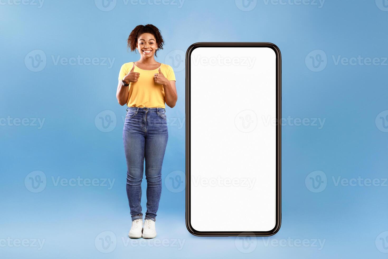 Best mobile offer. Smiling African American lady demonstrating giant smartphone with white screen and showing thumbs up with both hands on blue background, mockup copy space for app or website design photo
