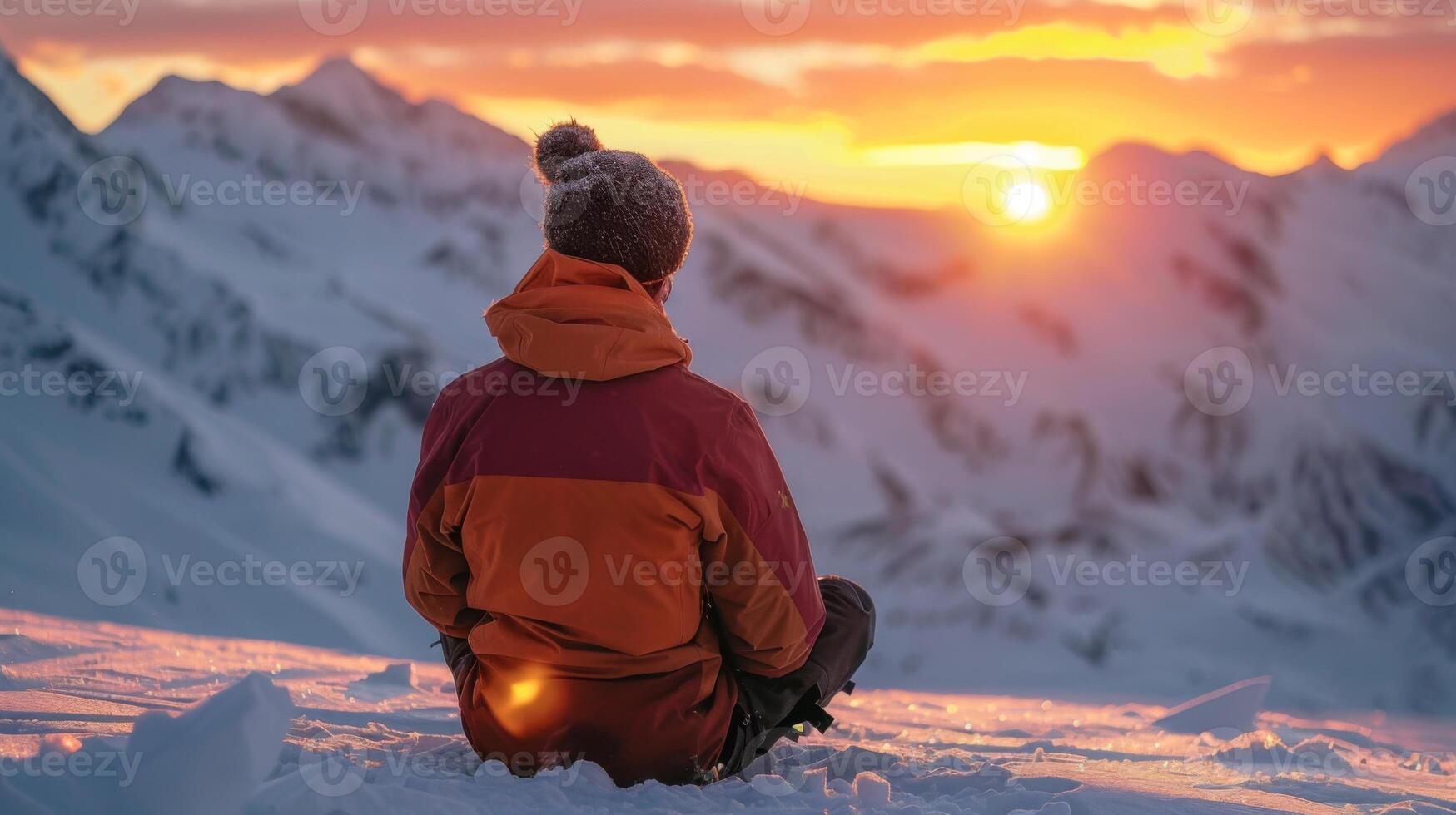 A person dressed in warm winter gear sits on snow, gazing at a vibrant sunset behind rugged mountain peaks, capturing the tranquility of the moment in a cold environment. photo