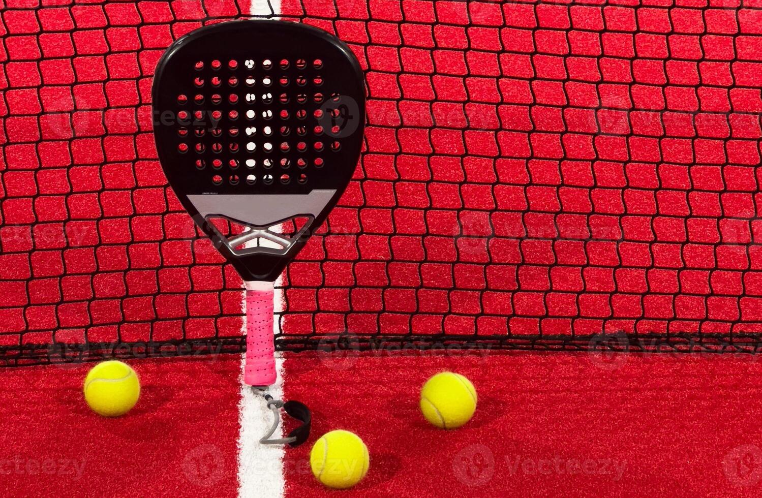 A padel or paddle tennis racket and a bright yellow tennis ball lie on a red court. The net photo