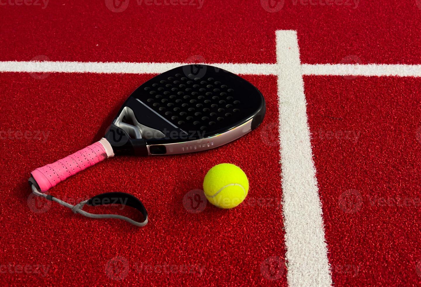 A padel or paddle tennis racket and a bright yellow tennis ball lie on a red court. The net photo