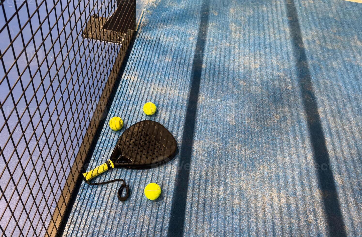 A padel or paddle tennis racket and a bright yellow tennis ball lie on a blue court. The net and a blurred figure of a player are visible in the background. photo
