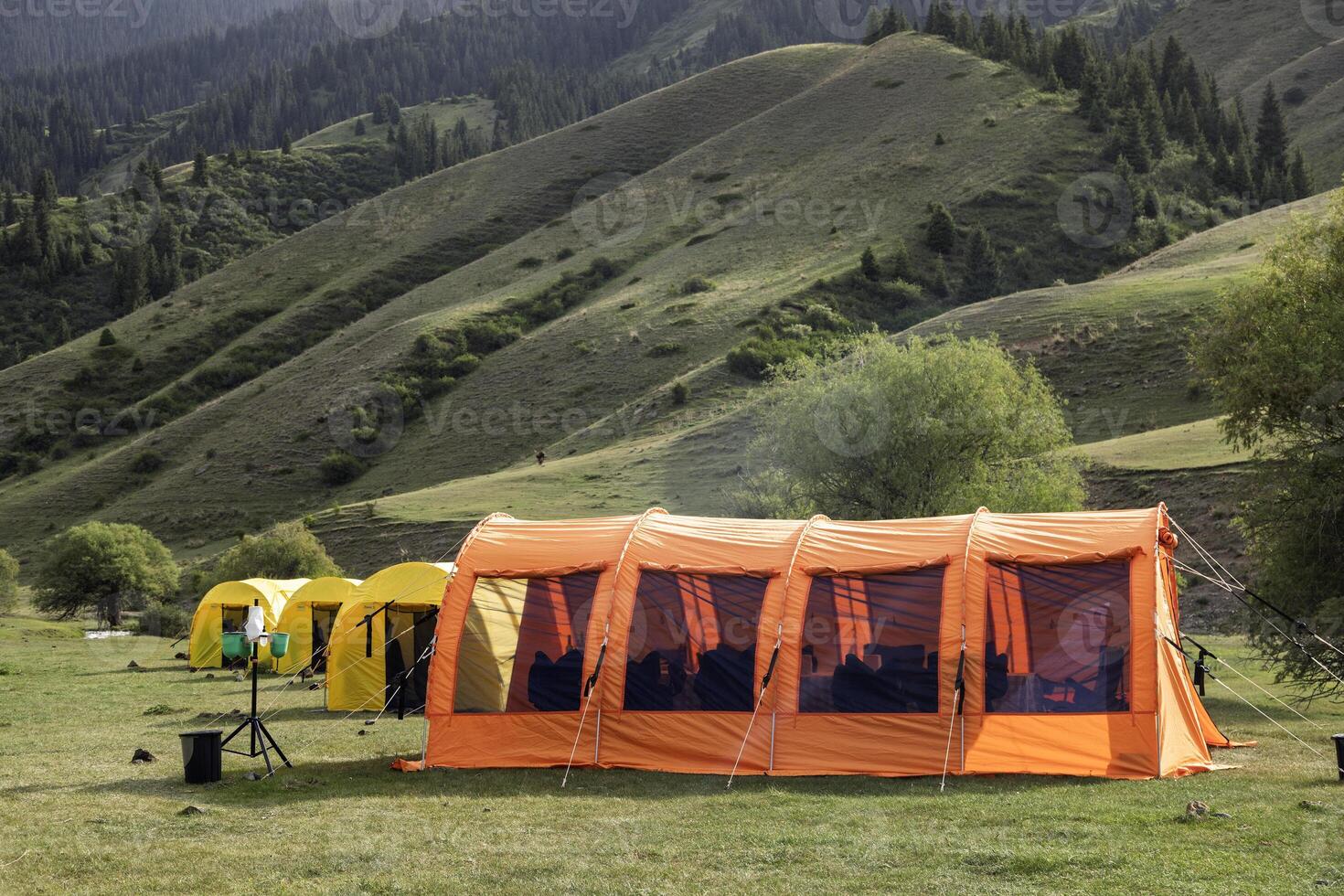 A large orange camping tent set in a scenic mountain valley, bathed in sunlight, surrounded by nature. photo