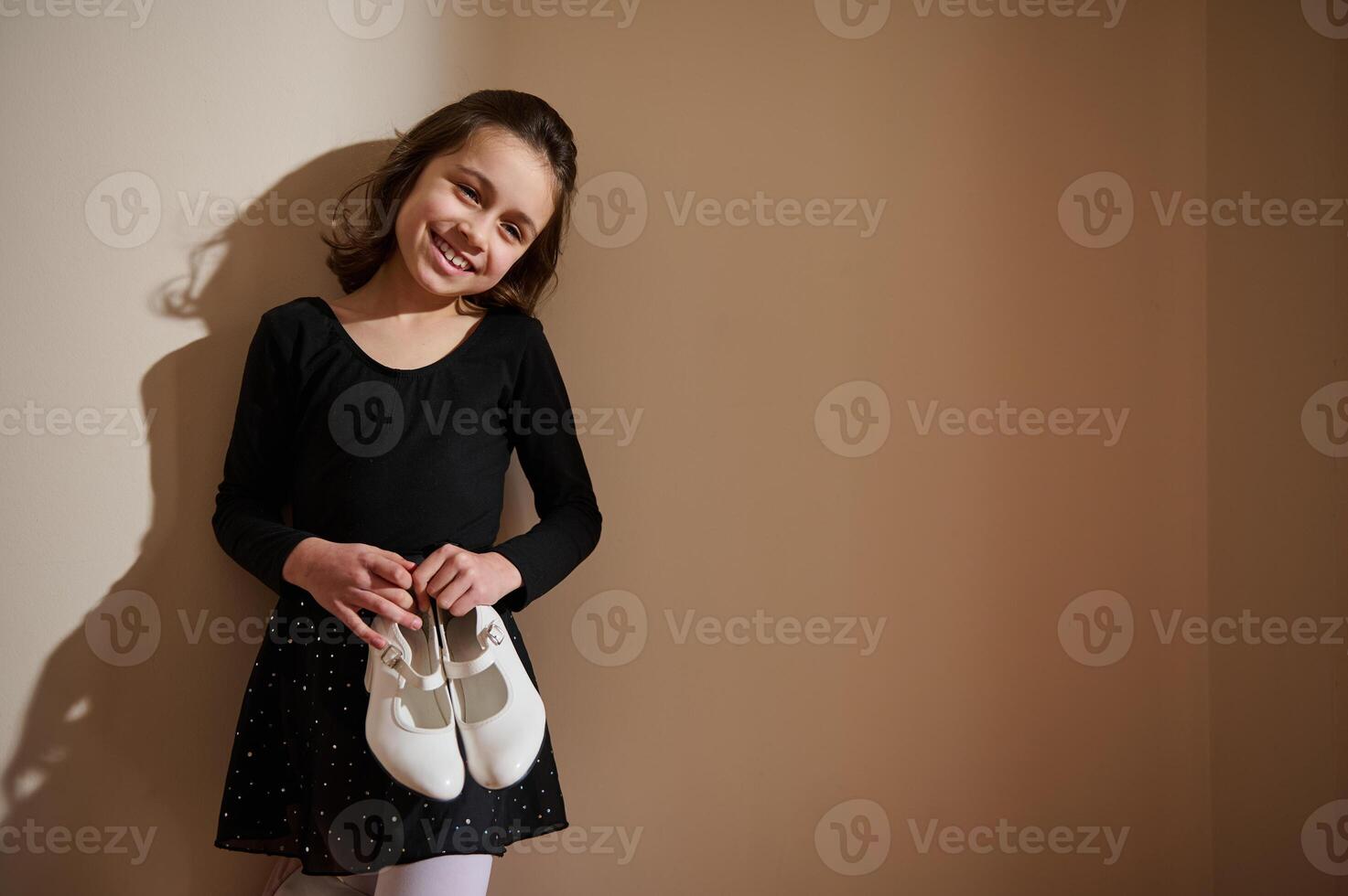 Smiling young girl in dance attire holding white ballet shoes indoors photo