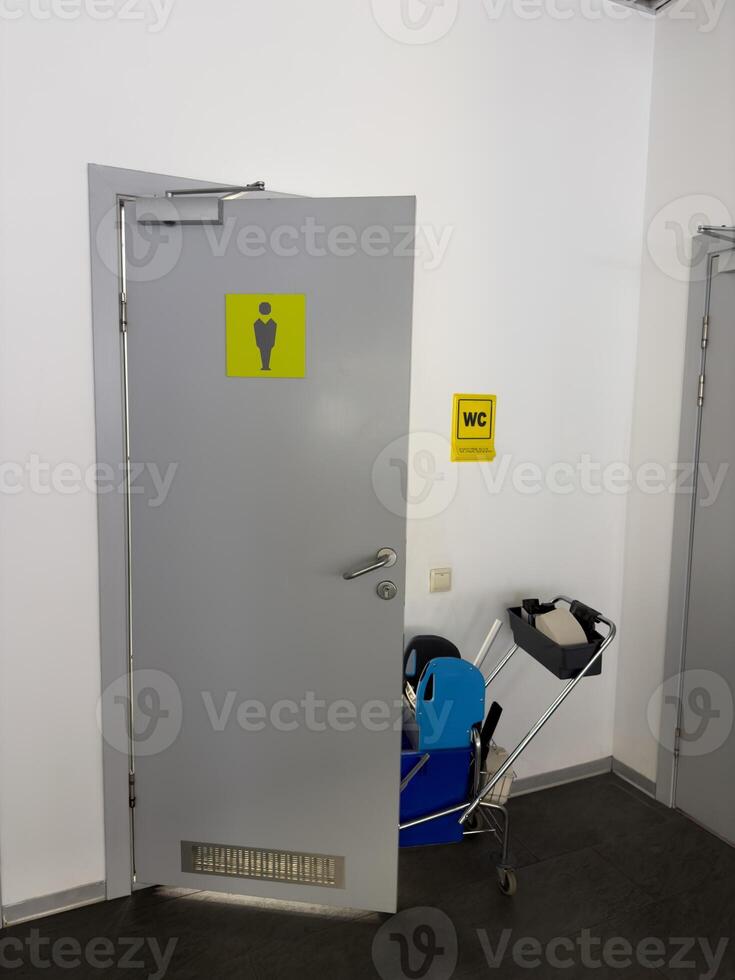 Door to men public toilet with cart for cleaning toilet service. Cleaning toilet in public place with help of cleaning trolley that serves wc, vertical photo