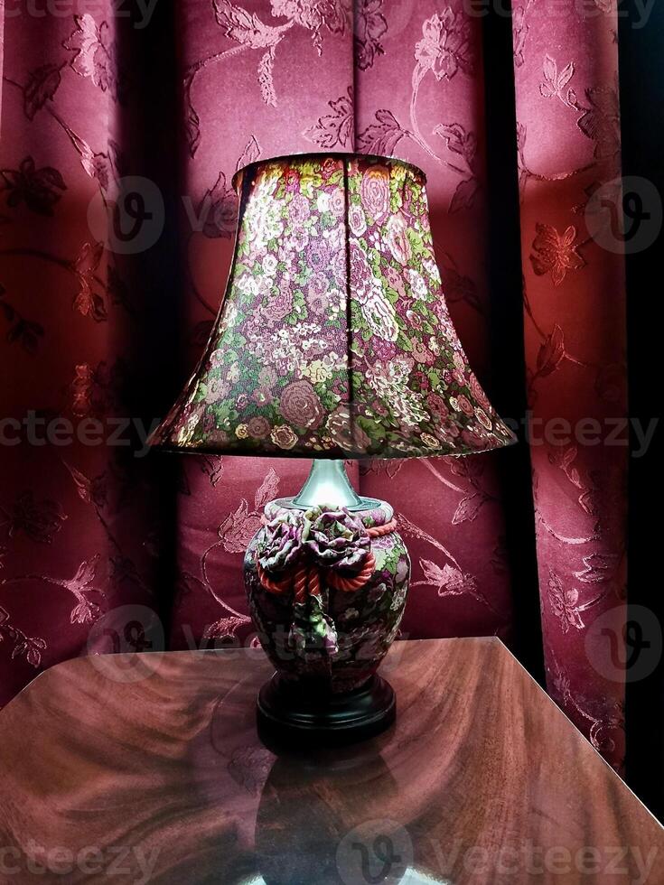 A beautifully crafted lamp is turned on, casting a warm glow in a room adorned with rich, floral-patterned curtains. It creates an inviting atmosphere photo
