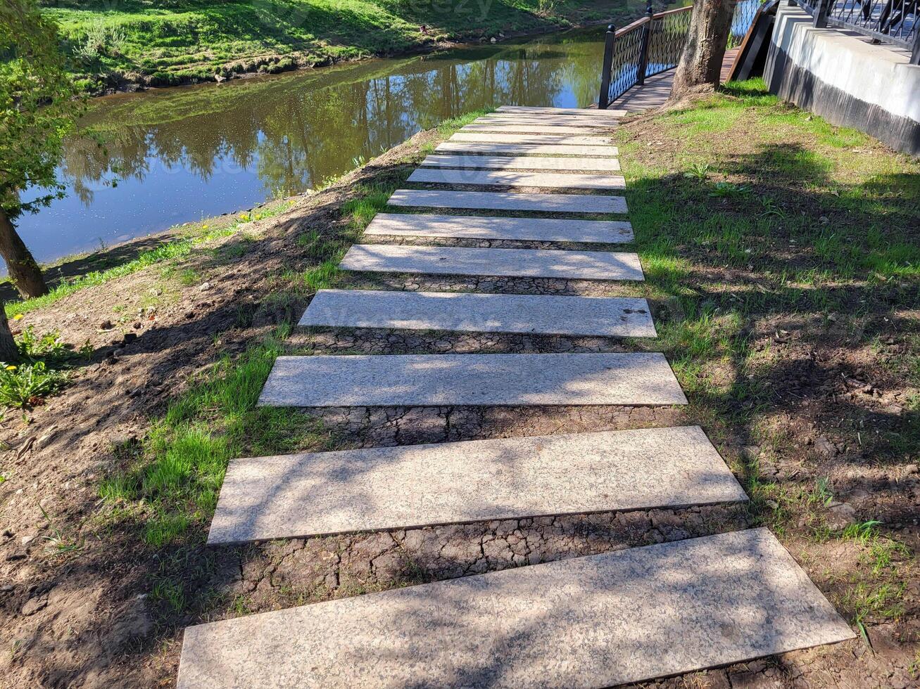 A path of concrete rectangles is laid along a shady spring lawn along the river. The concept of landscaping and landscape design photo