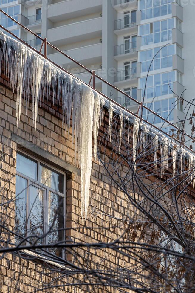 A group of sharp white transparent icicles is hanging down from the gray roof with ice of a building from orange bricks in winter. Vertical photo
