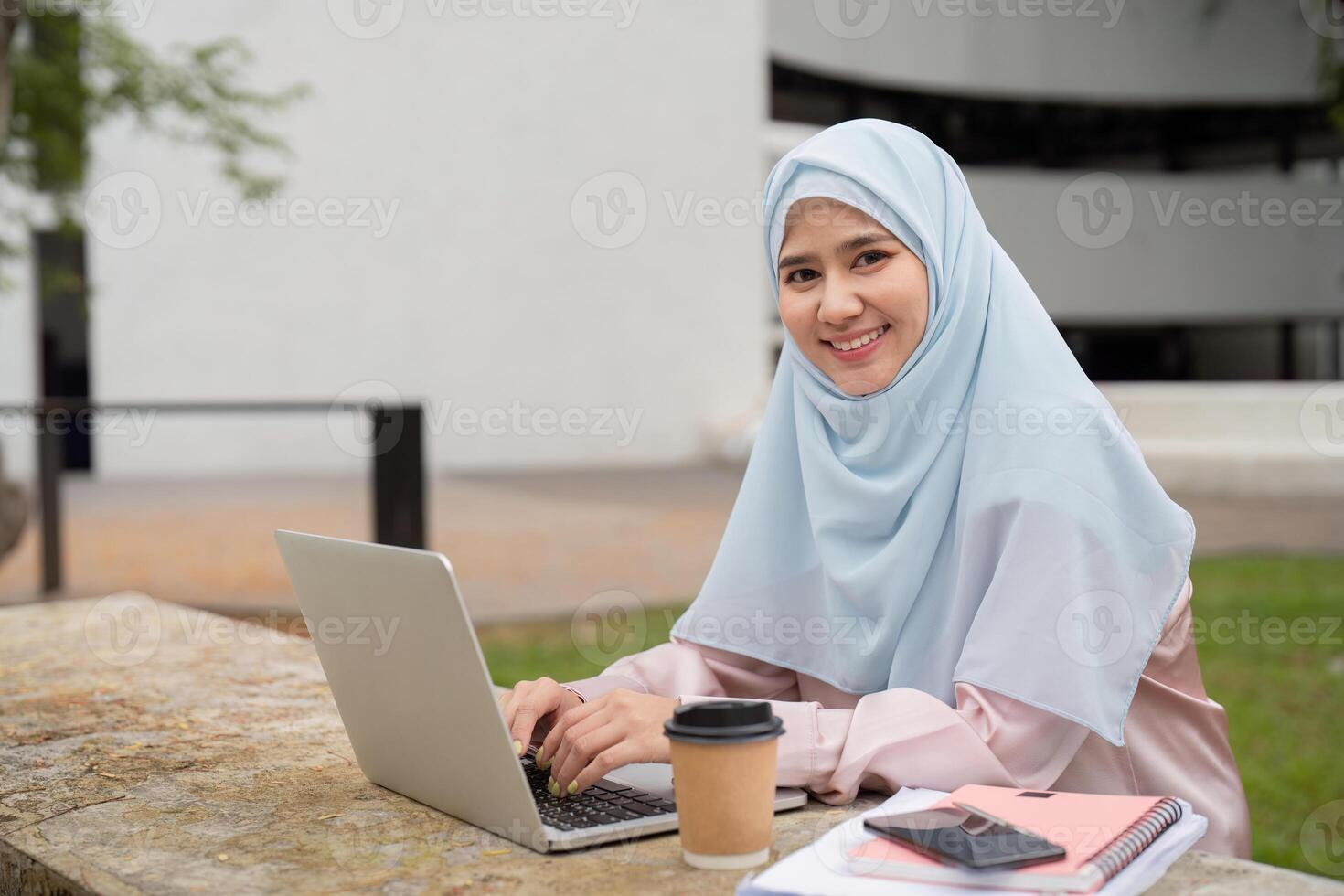 Smiling Muslim businesswoman in hijab working on a laptop with coffee outdoors. photo