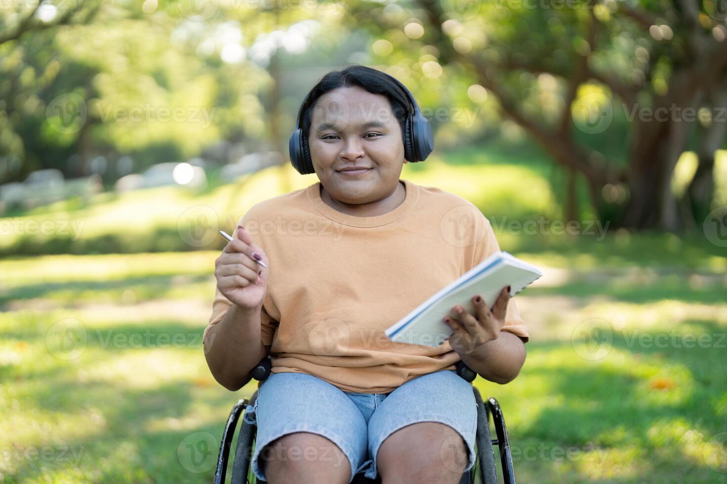 Diverse young man in wheelchair smiling while holding sketchpad and headphones photo