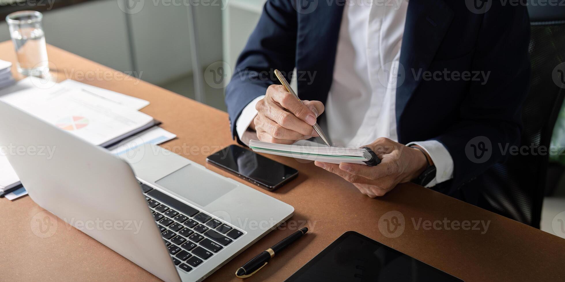 Elderly businessman taking notes in a notebook while working on a laptop at a modern office desk. photo