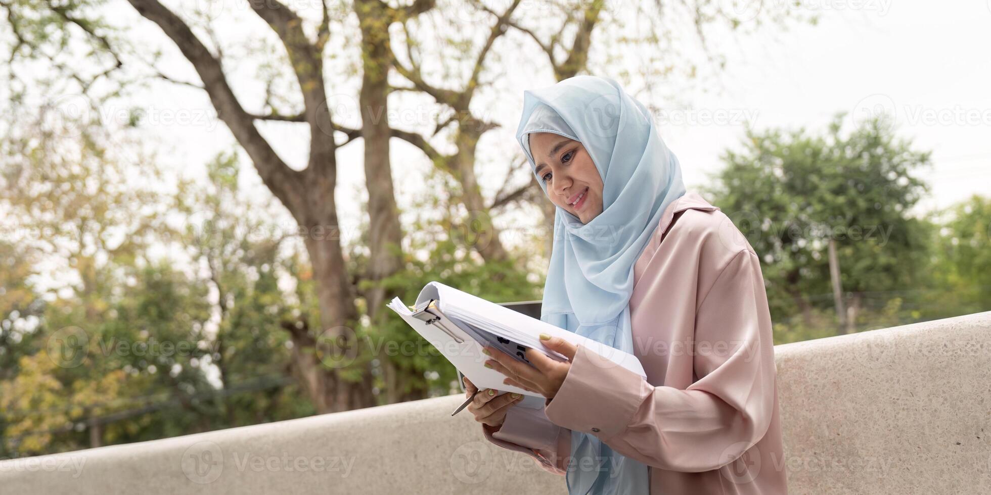 Smiling Muslim businesswoman reviewing documents in a serene outdoor environment. Showcasing dedication and cultural diversity in professional settings. photo