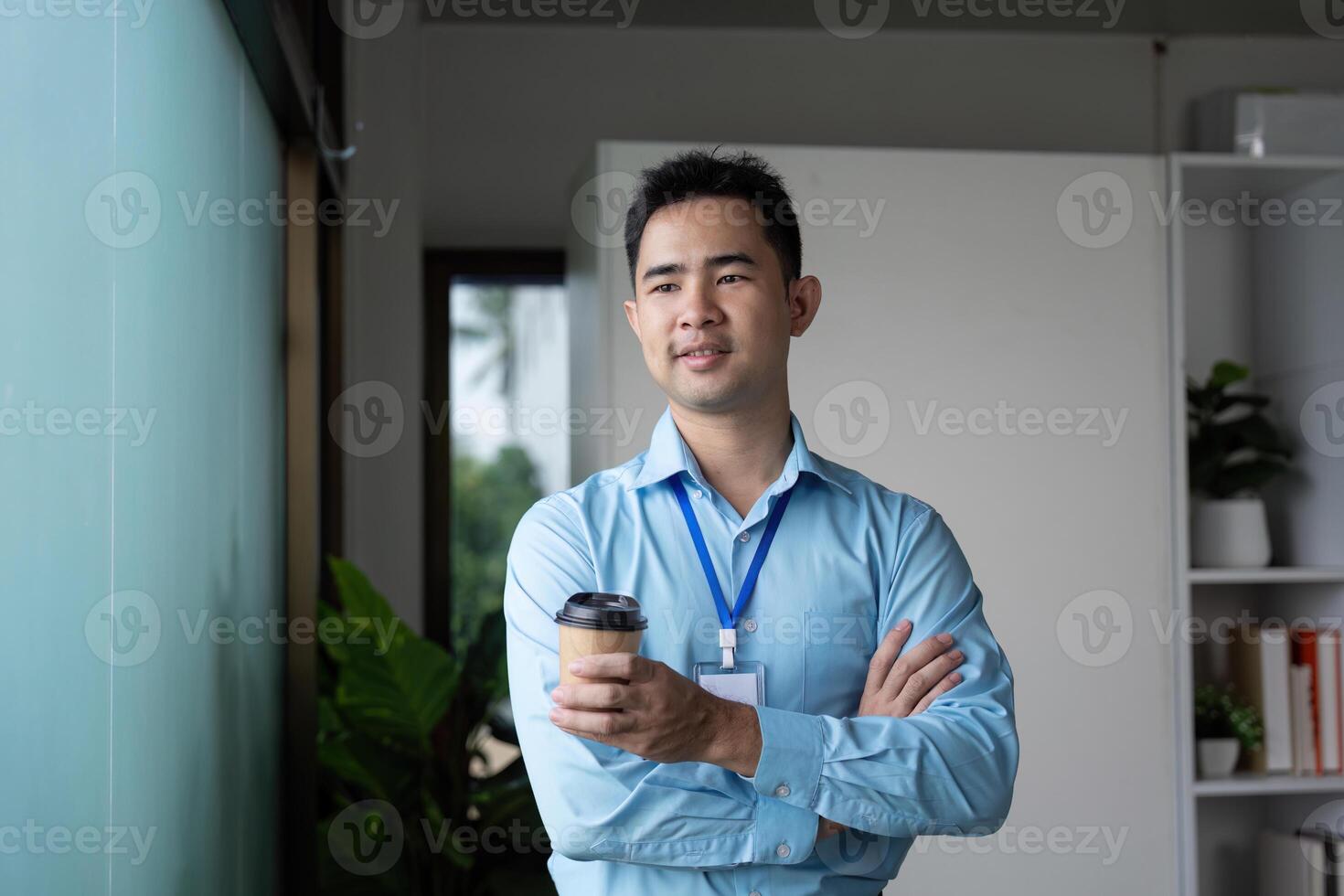 Confident Asian businessman holding a coffee cup, standing with arms crossed by a window in a modern office space. photo