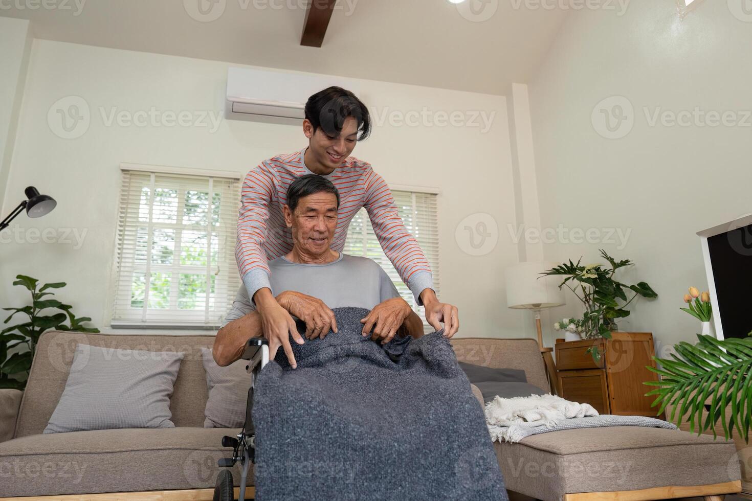 Son covering elderly father with a blanket while sitting in wheelchair at home. photo