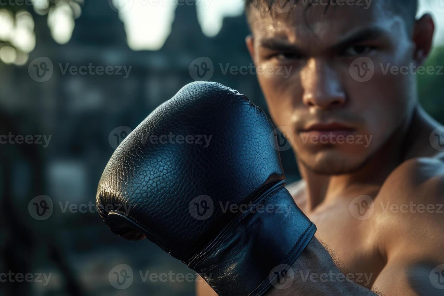 A Muay Thai fighter stands confidently, raising his gloved fist in preparation for practice. The backdrop features ancient temple ruins illuminated by the warm glow of sunset photo