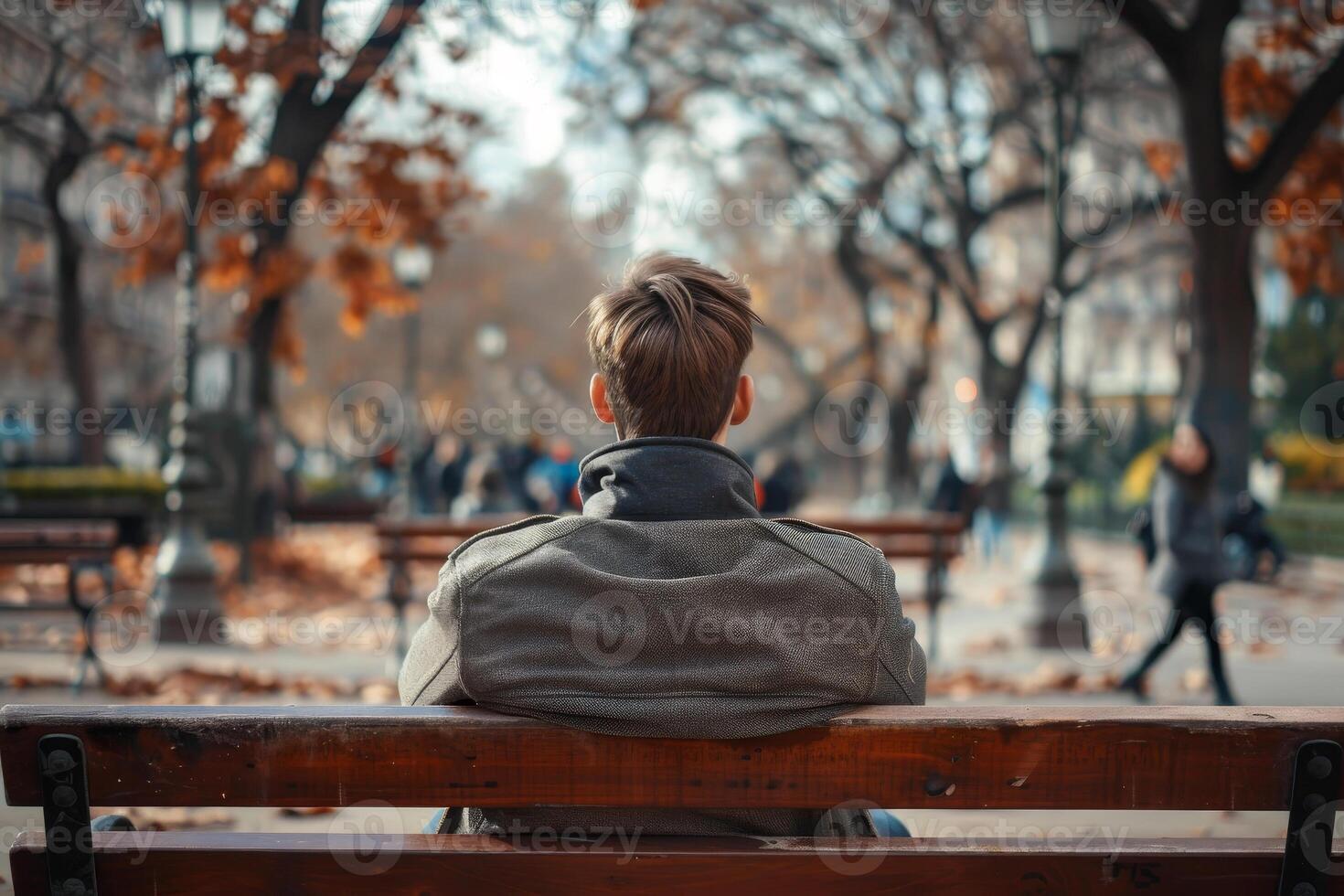 A person sits on a park bench, gazing thoughtfully into the distance as autumn leaves fall around them. Others stroll through the park, enjoying the crisp air photo