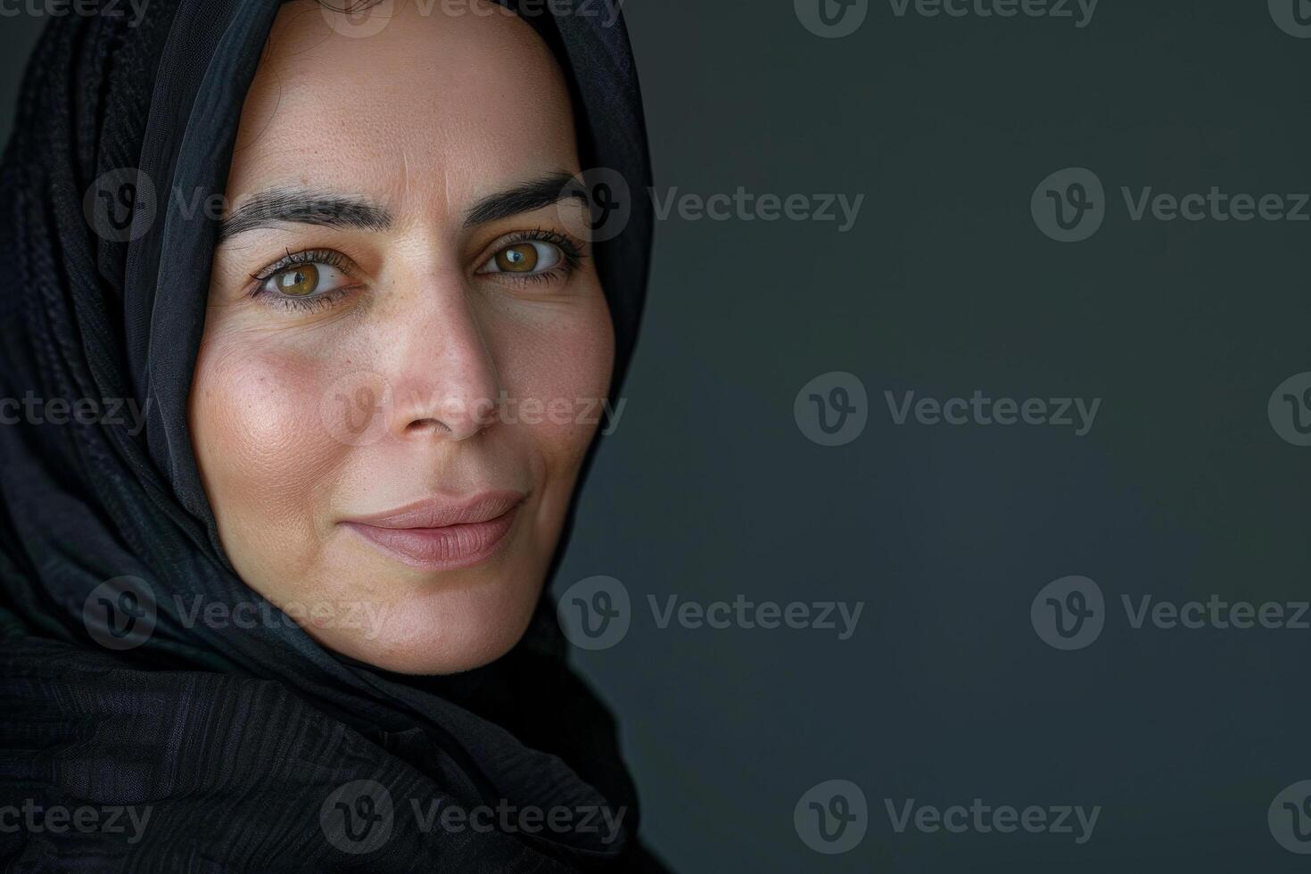 A woman with striking features and hazel eyes stands against a dark background, her black scarf framing her face as she looks directly ahead with a contemplative expression photo