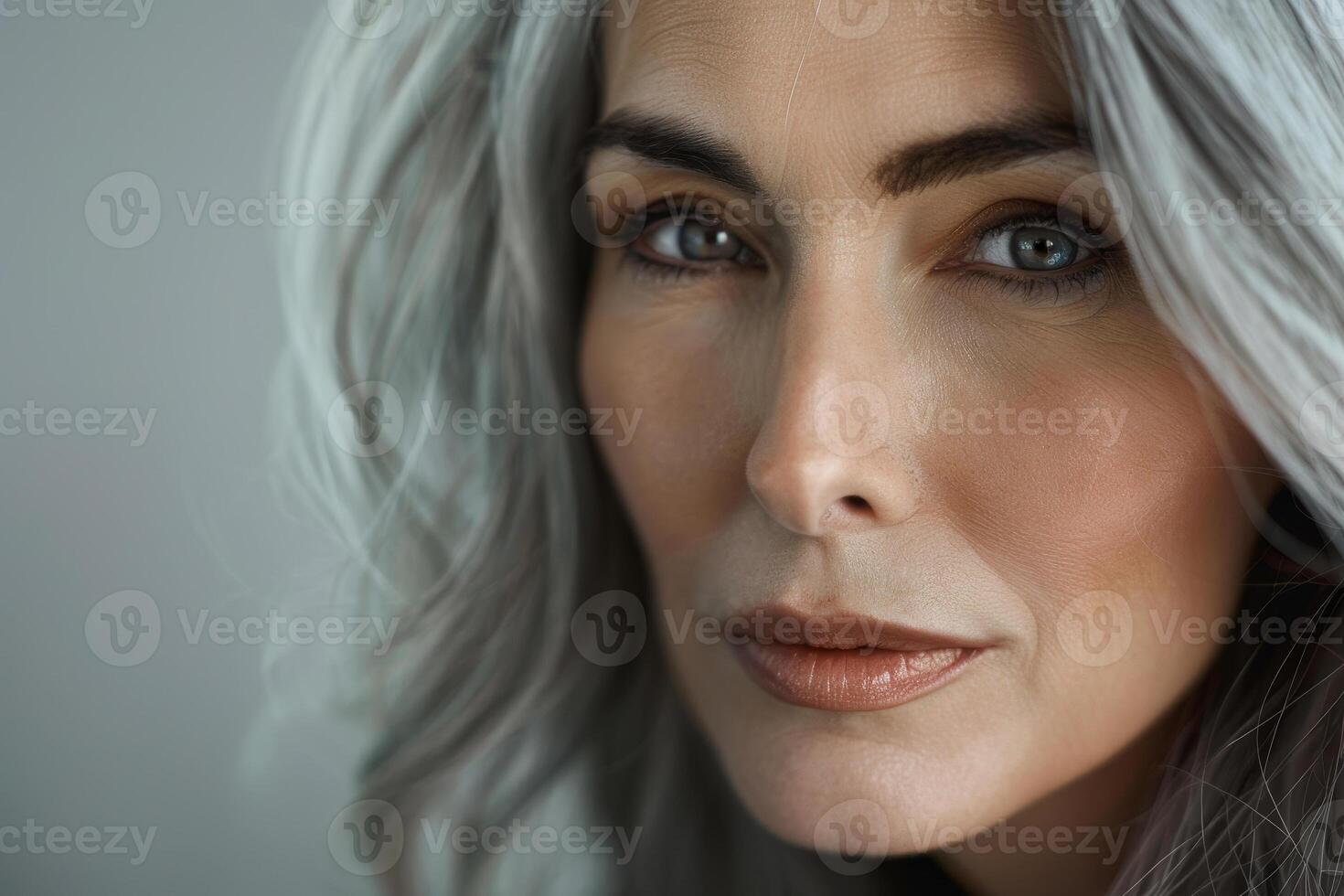 A woman with long gray hair and captivating blue eyes looks directly at the camera, radiating confidence and elegance with a subtle makeup look photo
