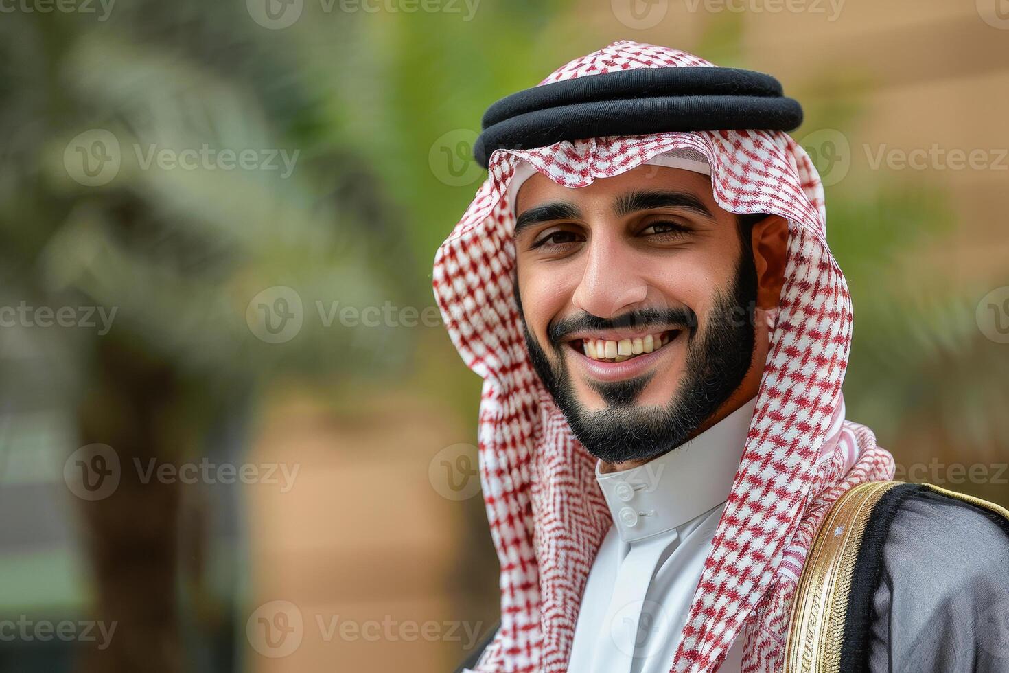 A young man dressed in traditional Saudi clothing poses for a portrait, showcasing a warm smile against a backdrop of palm trees and modern architecture photo