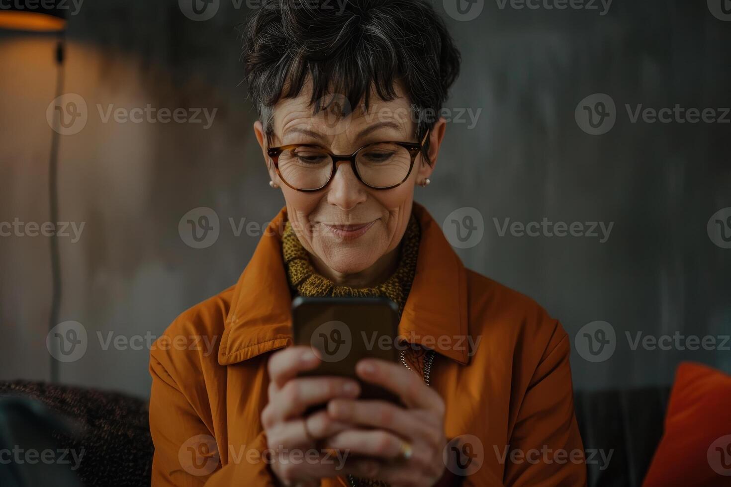 A woman in her sixties wearing a bright orange jacket enjoys using her smartphone, smiling softly as she engages with the device in a warm, inviting space photo
