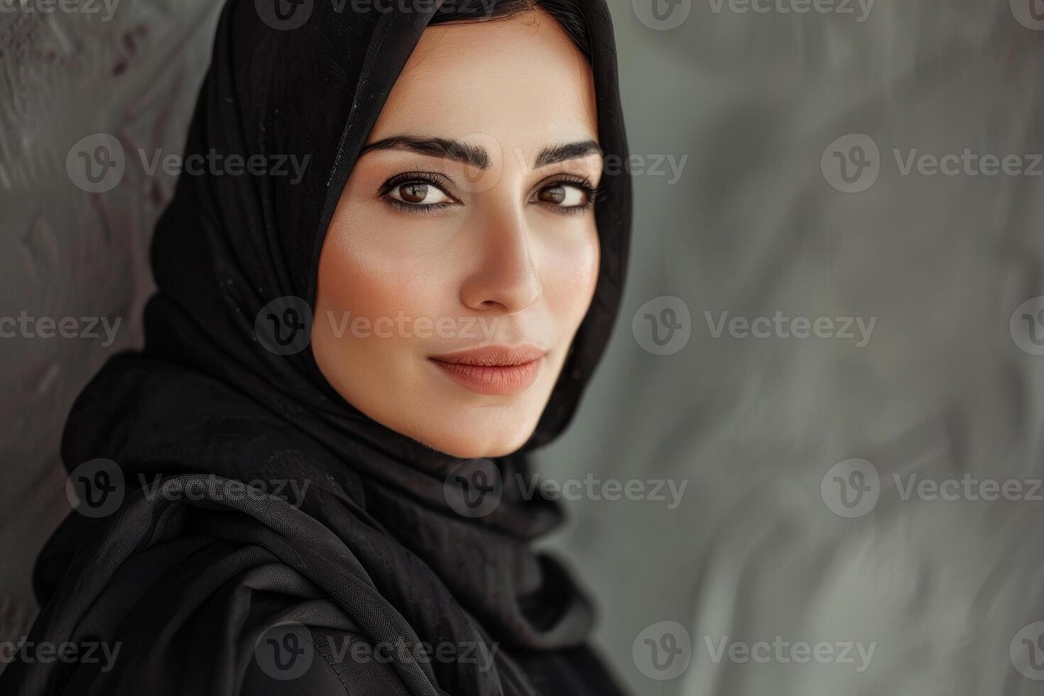 A woman wearing a black hijab gazes confidently towards the camera, framed by a softly blurred textured background. The warm light enhances her features photo