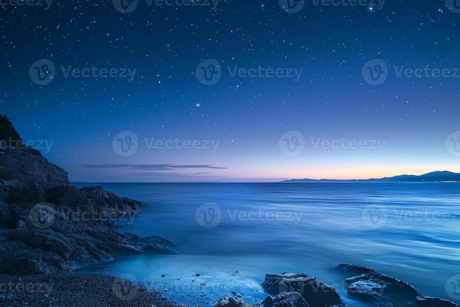 The sky transitions to twilight as stars emerge above a serene ocean. Gentle waves lap against the rocks, creating a peaceful atmosphere at dusk photo