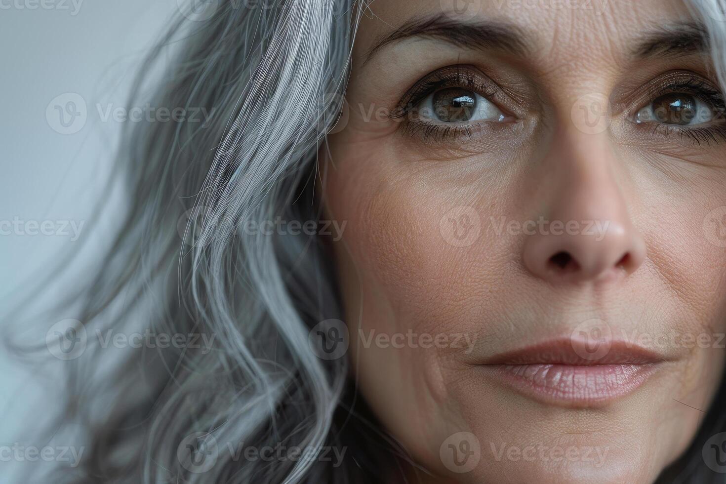 A mature woman with striking silver hair gazes thoughtfully, her expressive eyes reflecting wisdom and confidence. The soft indoor lighting highlights her features beautifully photo