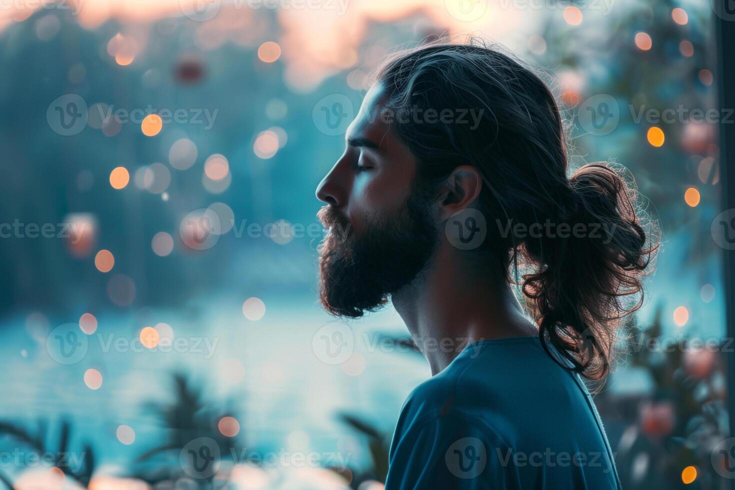A man with a beard and long hair is sitting in a room with a view of a lake photo