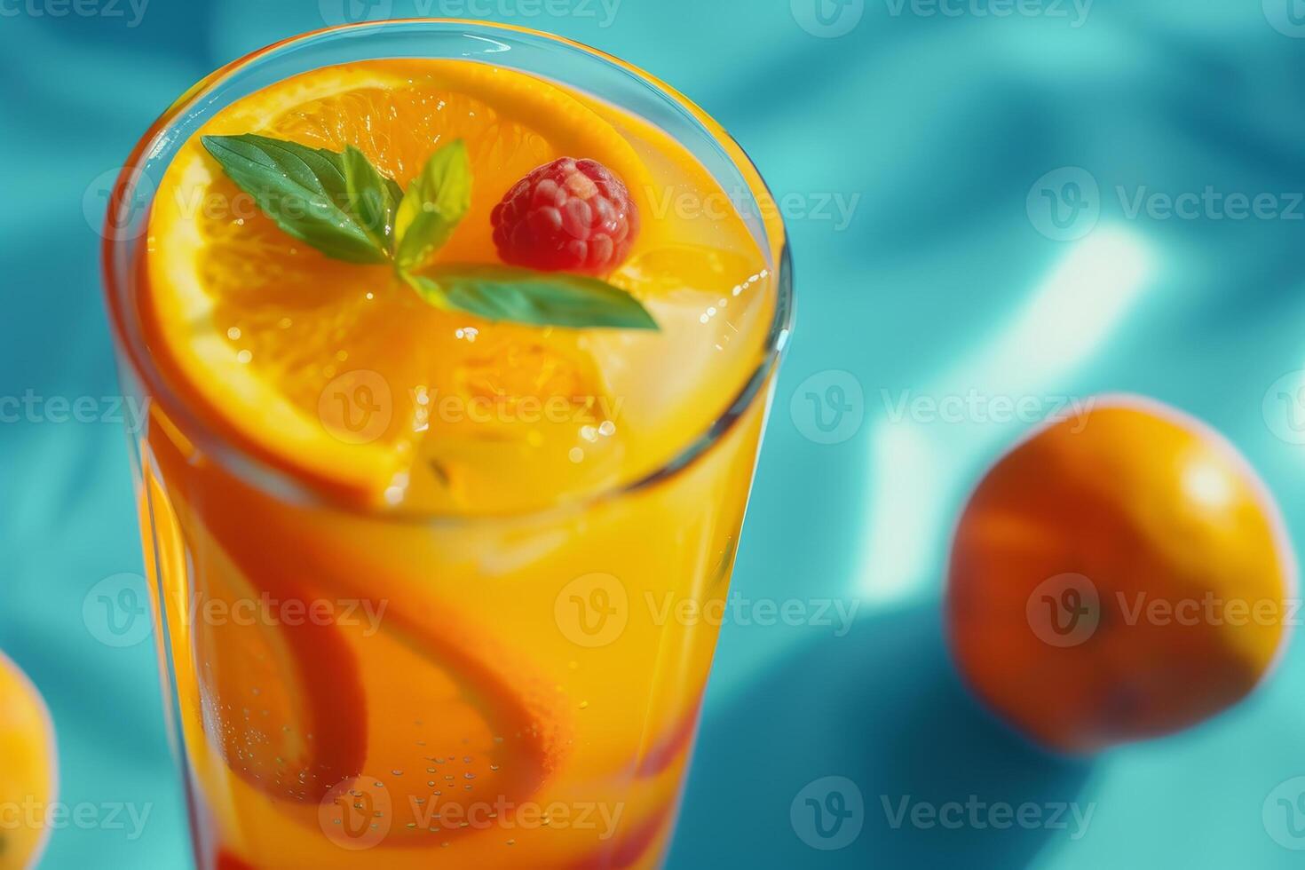 A tall glass filled with a colorful citrus beverage features orange slices, mint leaves, and raspberries, set against a bright blue backdrop with oranges nearby photo