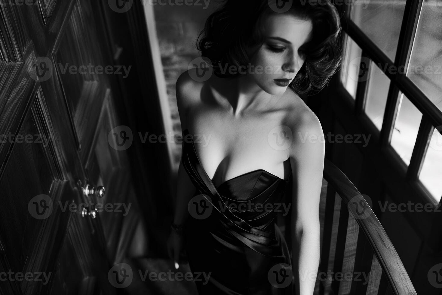 A woman in an elegant black dress stands gracefully on a staircase, her soft features illuminated by the warm glow of the hallway lights, creating a captivating atmosphere photo