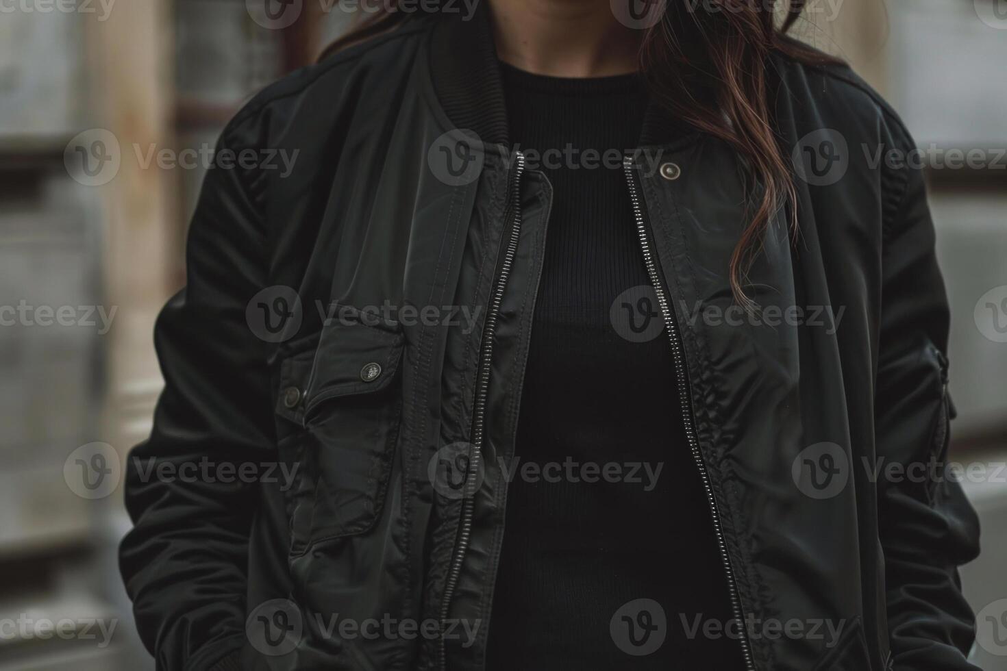 The individual is dressed in a fitted black bomber jacket paired with a black sweater, posing against an urban backdrop of subtle textures under an overcast sky photo