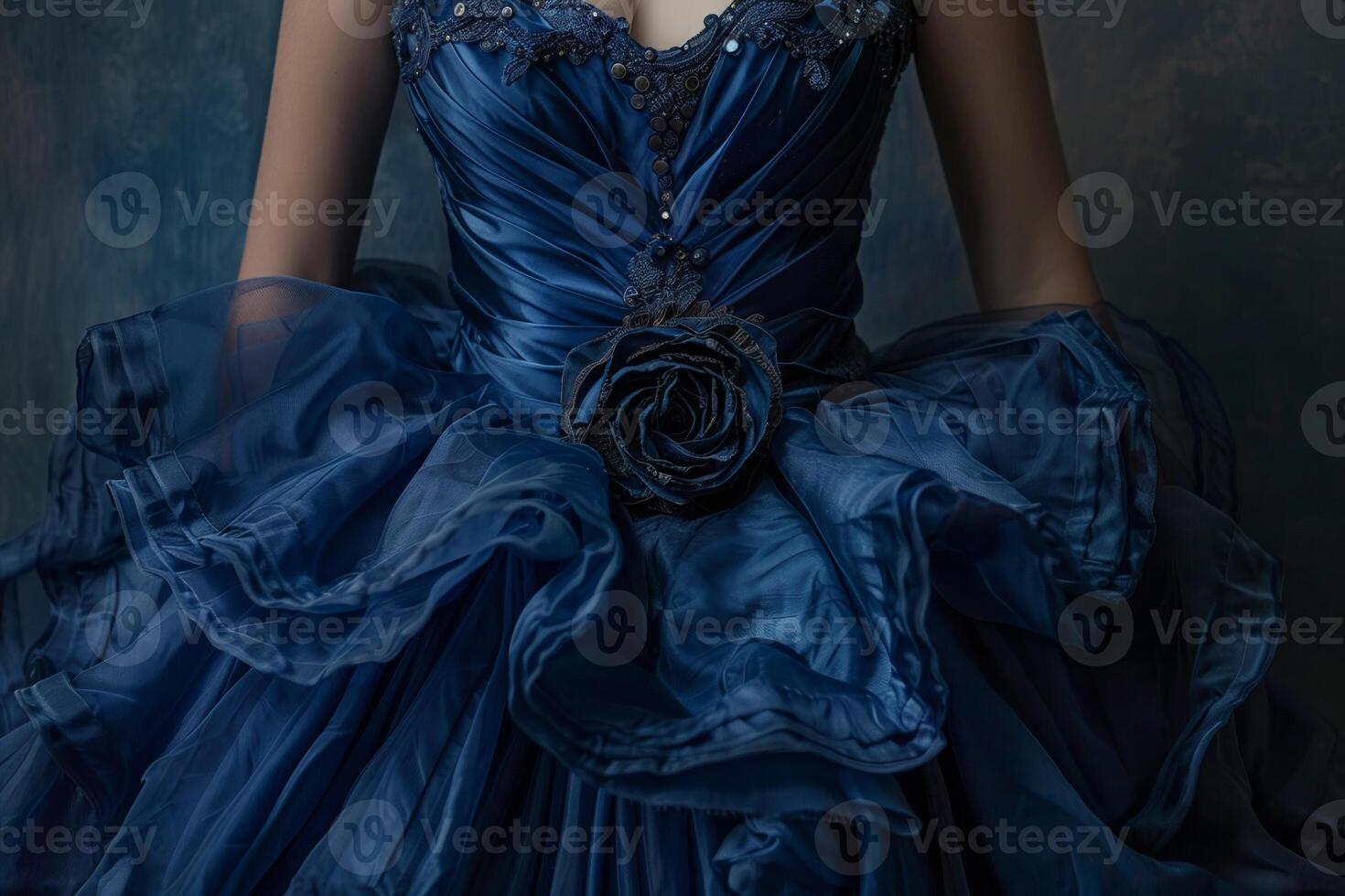 A person is wearing a stunning blue dress adorned with delicate embellishments, showcasing layers of flowing fabric and a rose accent. Soft lighting enhances the gown's elegance photo