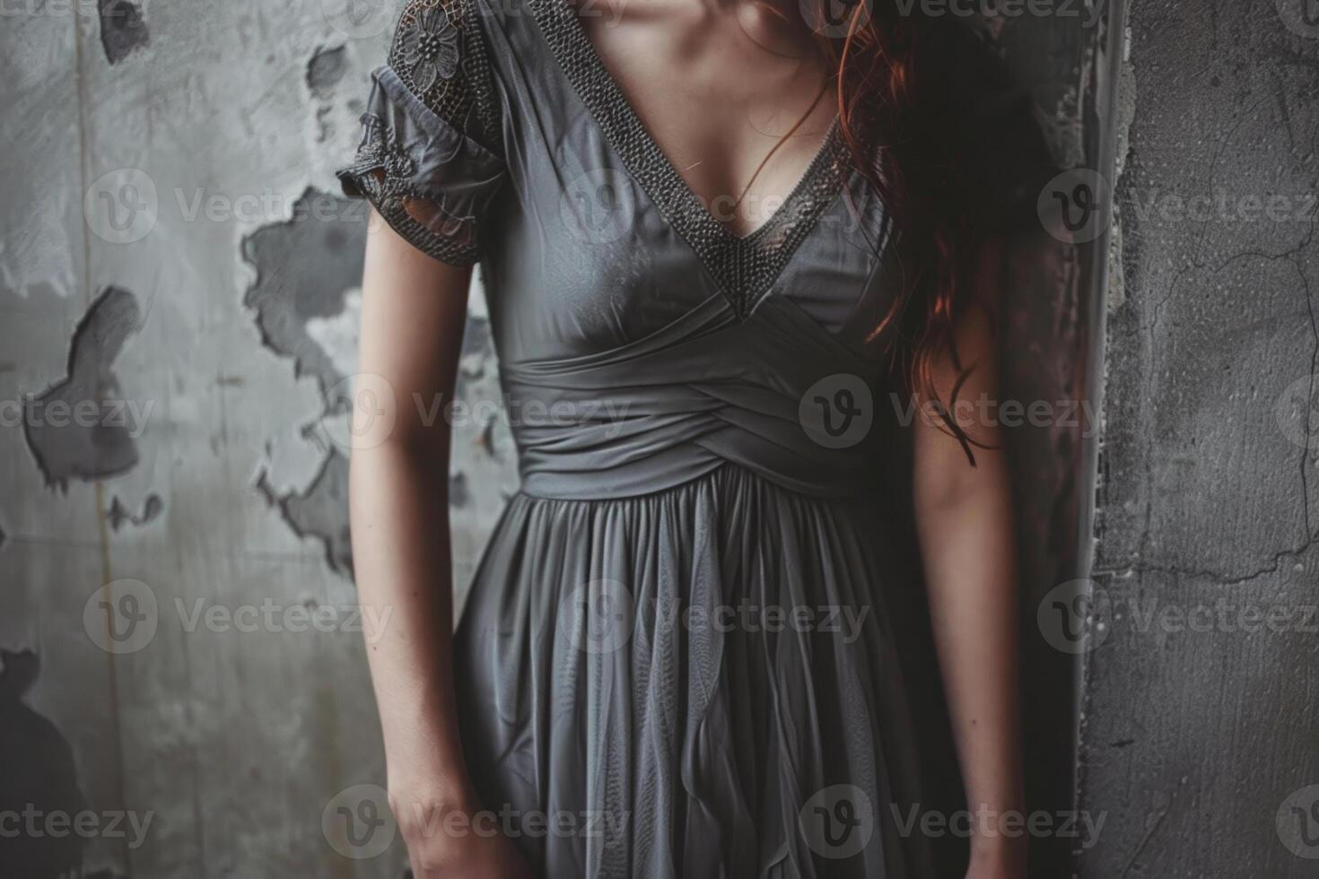 A young woman stands against a rough concrete wall, showcasing her gray dress with subtle details. The soft lighting highlights her features and the texture around her photo