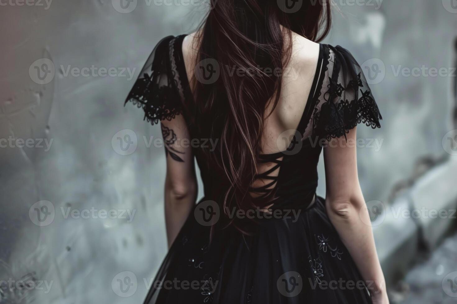 A woman wearing a black lace dress with delicate sleeves is positioned in front of a weathered gray wall. Her long hair cascades down her back, emphasizing the elegant design photo