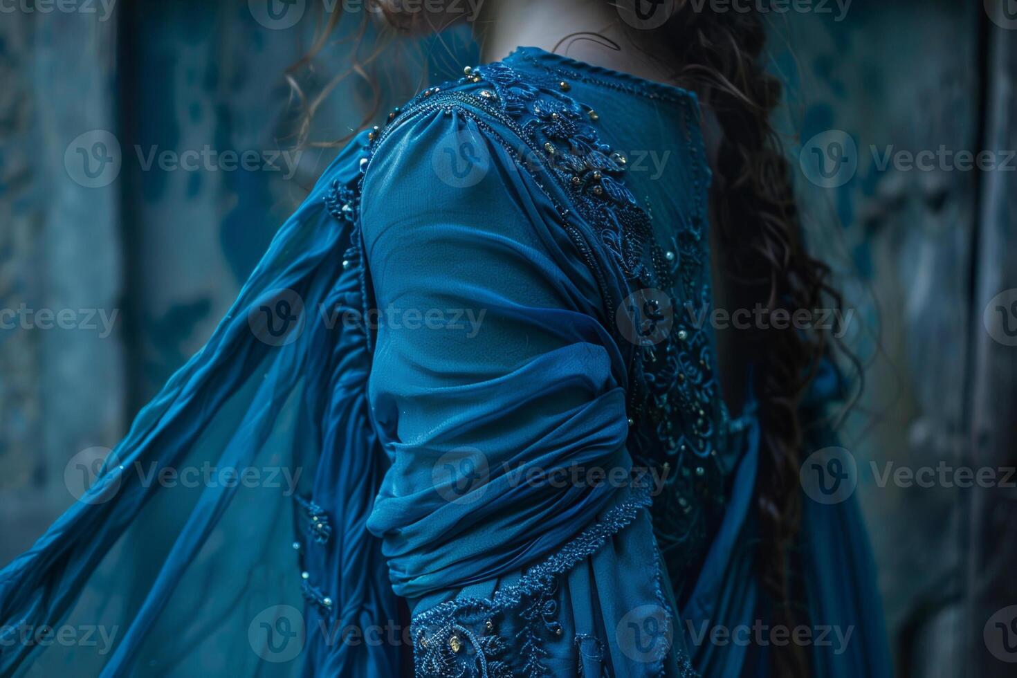A figure adorned in an intricate blue dress stands against a weathered wall, the flowing fabric gracefully catching the light, hinting at a moment of serene reflection photo
