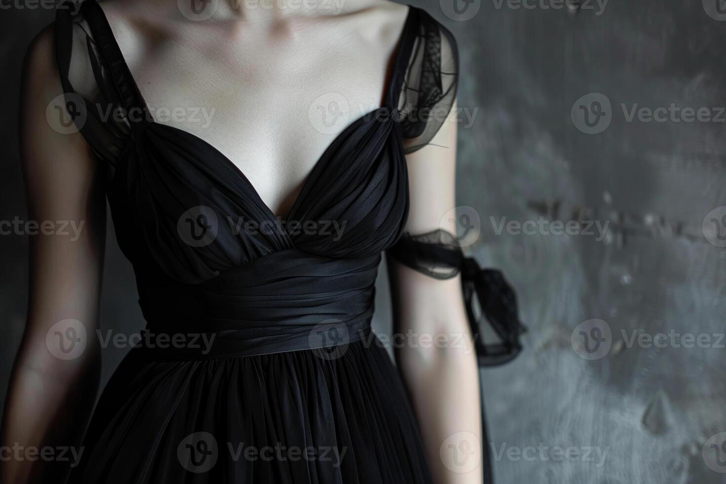 A woman in an elegant black dress with delicate sheer straps stands against a textured gray backdrop, emphasizing the garment's flowing design and fabric details photo