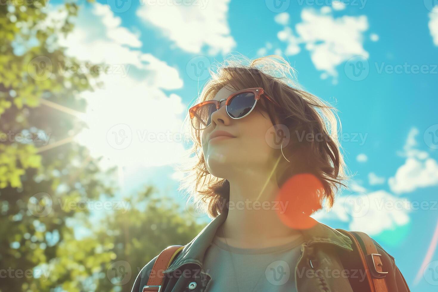 A woman wearing sunglasses and a backpack is looking up at the sky photo